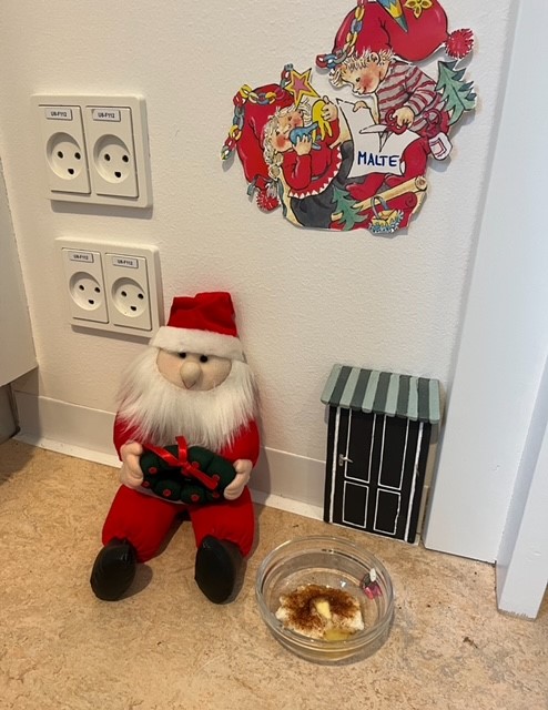 Julemandspyntet dukke siddende på gulv ved stikkontakter, med skål med mad foran. Bagpå en farverig julegalleri på væggen