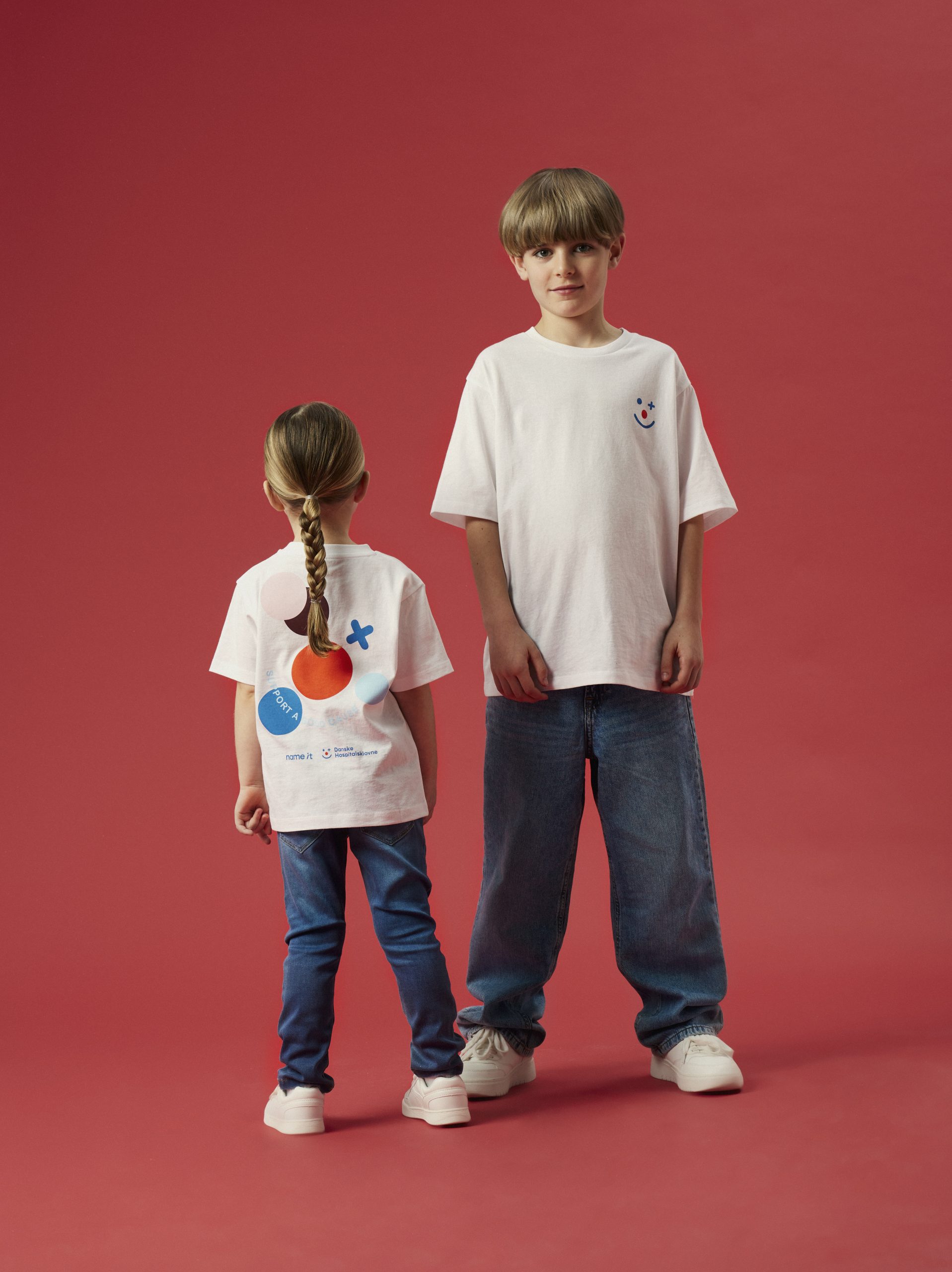 To børn stående foran en rød baggrund iført hvide t-shirts med Danske Hospitalsklovne-logoet; det ene barn ses forfra, det andet med ryggen til, så logoet på ryggen er synligt
