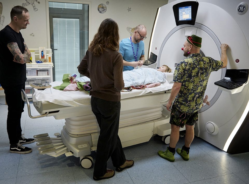 En radiograf, en hospitalsklovn og en kvinde står foran en MR-scanner, hvor der ligger en pige i