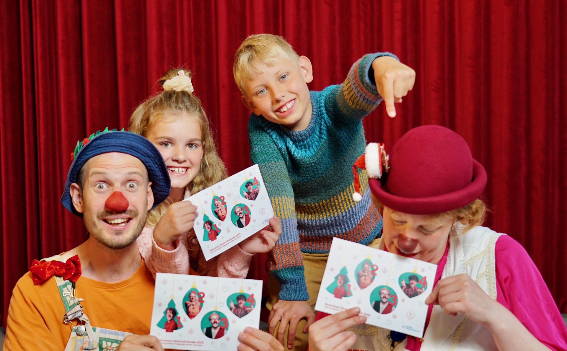 To små børn, en dreng og en pige, sidder sammen med de to hospitalsklovne, Diamanda og Bob, og fremviser årets adventskalender fra Julelotteriet.
